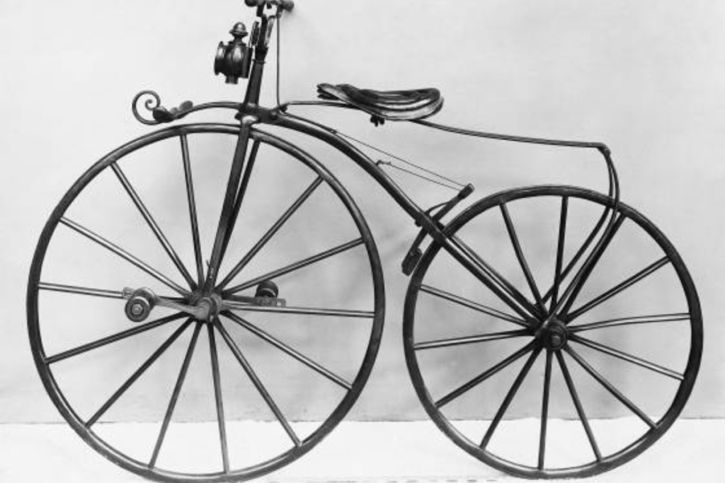A imagem em preto e branco apresenta uma bicicleta clássica estilo 1870, uma das primeiras bicicletas já criadas. A bicicleta é alta e desengonçada, com rodas grandes e um quadro em forma de 