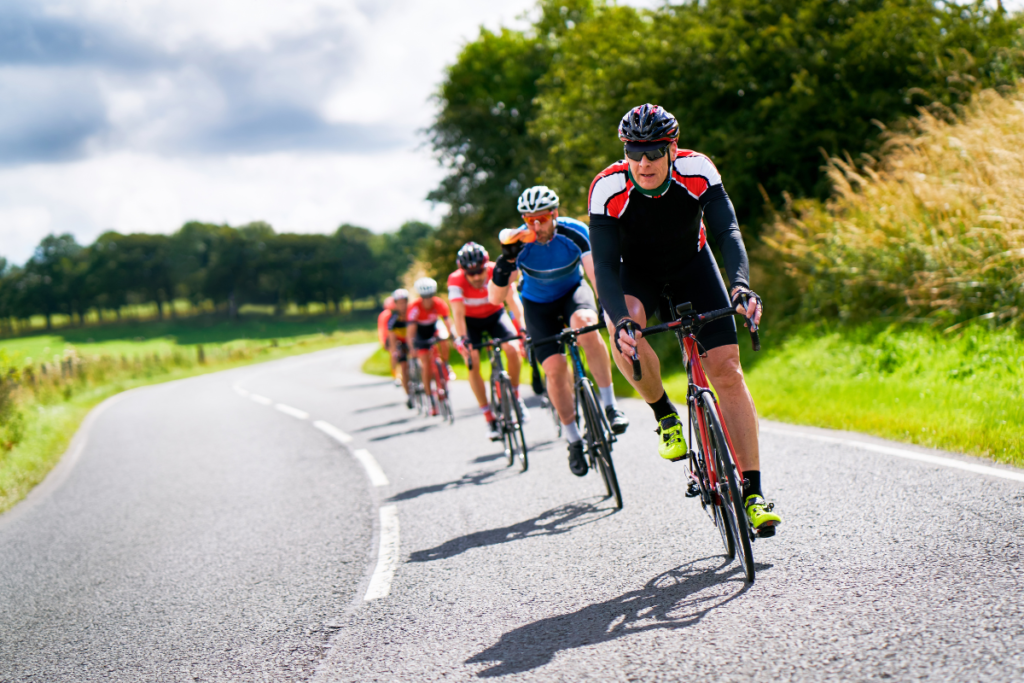 A imagem mostra um grupo de ciclistas descendo uma estrada em formação. Eles estão todos vestidos com roupas de ciclismo, capacetes e óculos de sol. A estrada é íngreme e sinuosa, com algumas curvas acentuadas e alguns trechos retos.