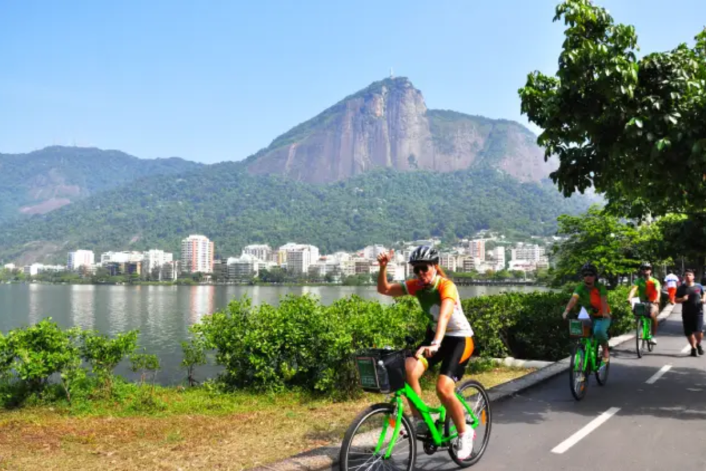 Na imagem, podemos ver uma mulher pedalando em uma bicicleta de corrida pela ciclovia da Lagoa Rodrigo de Freitas, no Rio de Janeiro. Ela está usando uma roupa de ciclismo justa ao corpo, com cores vibrantes e um capacete de segurança para proteger sua cabeça.
