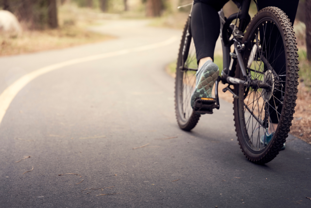 A imagem retrata uma mulher que está parada em sua bicicleta, que é preta, na estrada. A mulher está vestindo uma roupa preta apropriada para ciclismo, com shorts ajustados e uma camisa de tecido respirável. A postura da mulher na bicicleta sugere que ela está confiante e confortável em sua bicicleta, pronta para seguir em frente a qualquer momento.
