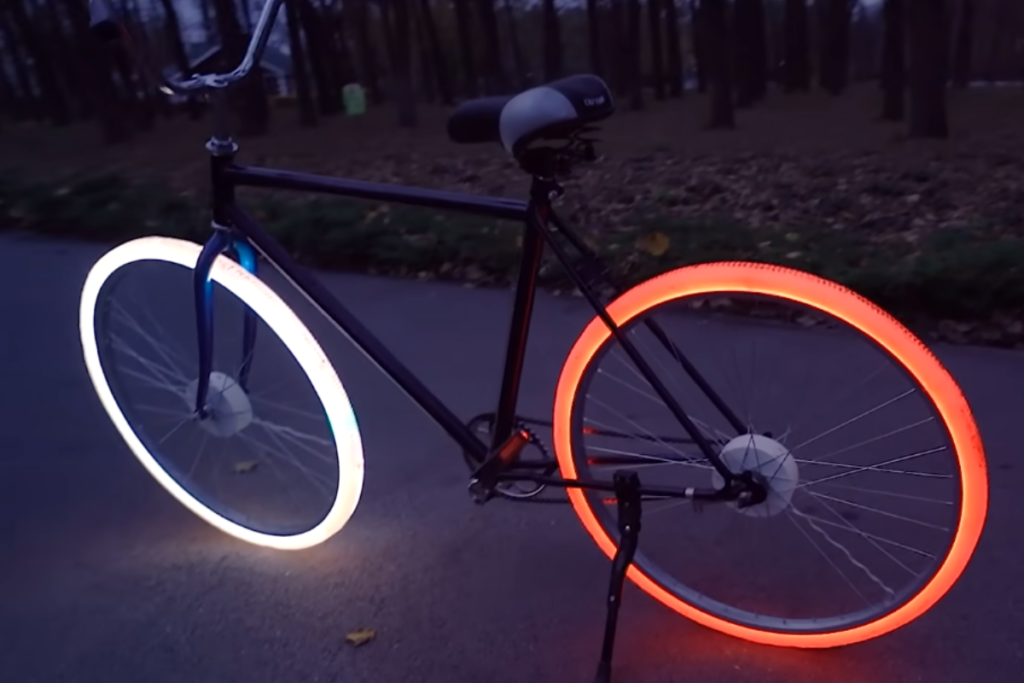 Nesta imagem, vemos uma bicicleta impressionante e futurista com rodas equipadas com luzes de LED. A roda traseira emite uma luz vermelha vibrante, enquanto a roda dianteira brilha em um branco deslumbrante.